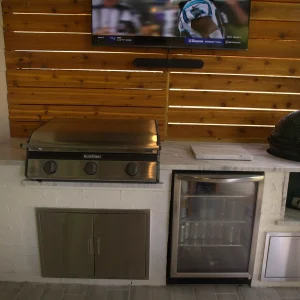 An outdoor kitchen setup featuring a stainless steel grill, a mini fridge stocked with drinks, a green ceramic cooker, and a TV mounted above on a wooden privacy wall.