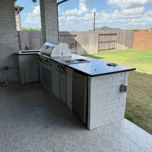 Outdoor kitchen with a built-in grill, sink, and counter space set against a white brick wall on a covered patio. Patterned tile flooring and a backyard with a wooden fence are visible.