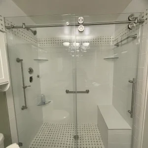 A modern glass-enclosed shower with sliding doors, white tile walls, a built-in bench, grab bars, two corner shelves, and patterned floor tiles—another elegant space by your trusted Kitchen Remodeling Company in Houston. Paper towel rolls are visible in the foreground left.