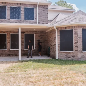 Two people stand under a covered patio at the back of a brick house. The house windows are covered with dark screens. The lawn in the foreground is partly brown and dry.