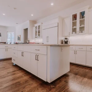 Spacious modern kitchen with white cabinets, an island in the center, stainless steel appliances, hardwood floors, and large windows allowing natural light to fill the room.