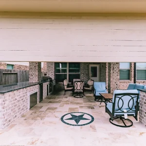A covered backyard patio with tile flooring, an outdoor kitchen on the left, cushioned seating around tables, and a large star design on the floor, surrounded by brick walls and a fence.