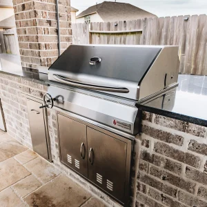 A built-in stainless steel outdoor grill is set into a brick patio countertop, featuring a closed lid, control knob, and double-door storage below, with a fenced backyard visible in the background.