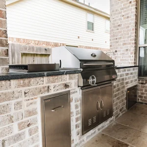A built-in stainless steel grill and storage cabinets are set in a brick outdoor kitchen on a patio, with a house and wooden fence visible in the background.