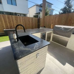 Sleek Outdoor Kitchen featuring a black countertop, built-in sink, stainless steel appliances, covered grill, and light brick base. Wooden fencing and contemporary houses provide a stylish backdrop under a clear sky.