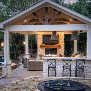 Covered outdoor patio with a stone bar, three barstools, a built-in fireplace, TV, sofa, and hanging lights. Surrounded by trees, a pool is on the left, and a black fire pit is in the foreground.
