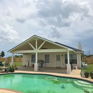A backyard with a covered patio attached to a house, featuring outdoor furniture, potted plants, and a swimming pool in the foreground. The sky is cloudy and a wooden fence surrounds the yard.