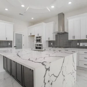 Modern kitchen with white cabinets, gray backsplash, stainless steel appliances, and a large island with a white marble countertop featuring bold dark veining. Recessed lighting brightens the clean, open space.