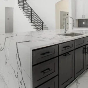 Modern kitchen with a large marble island featuring dark cabinets, a built-in sink, and stainless steel appliances. The space has white marble floors, white walls, and a black staircase in the background.